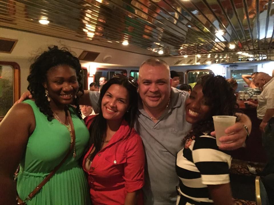 People on a boat deck at night with pink lights, facing a city skyline across the water.