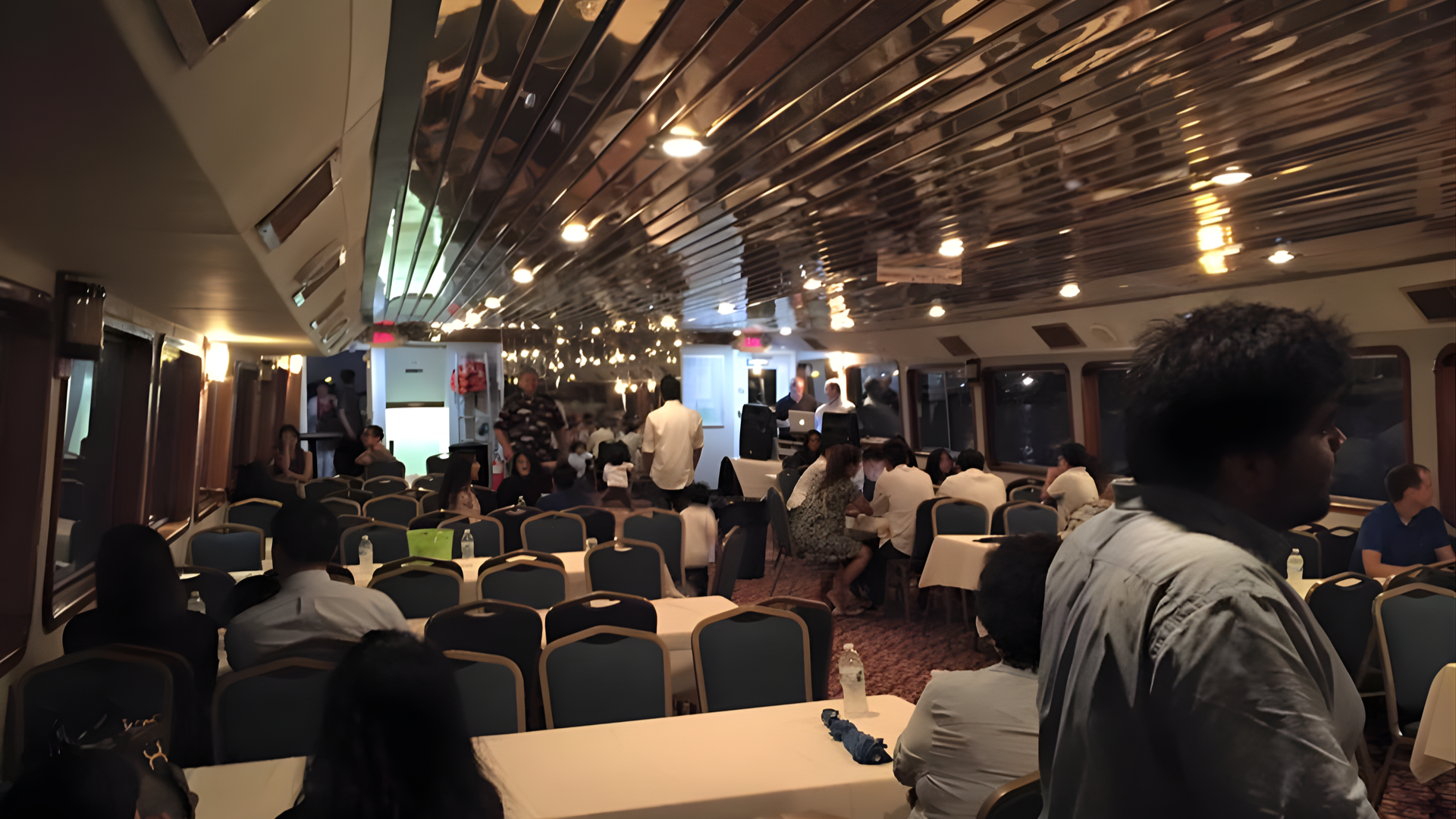 Crowded indoor event hall with seated attendees, warm lighting, and people standing near a stage at the front