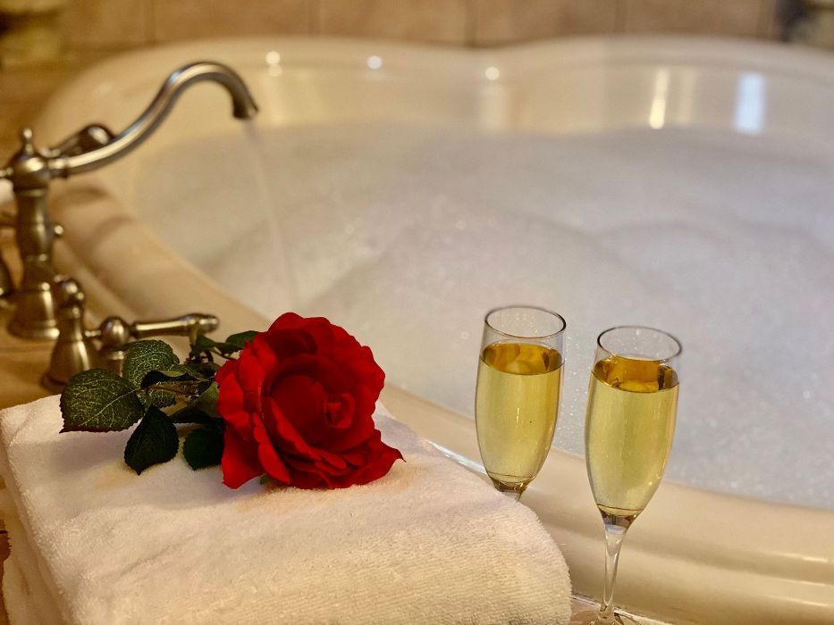 Heart-shaped bathtub filled with bubbles, two champagne flutes, red rose, and folded white towels, romantic spa setting.