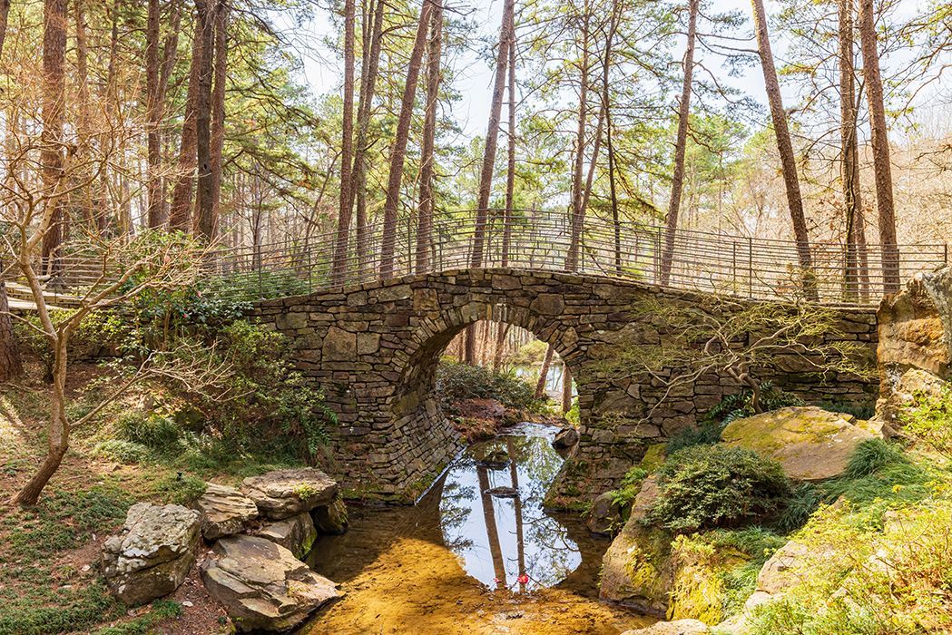 Stone arch bridge over a stream, surrounded by trees in a wooded area.