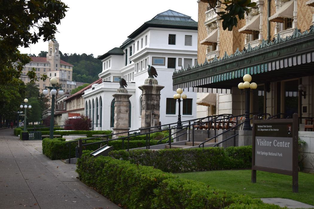 Street view of bathhouses and gardens in Hot Springs, Arkansas. White buildings, greenery, and a cloudy sky.