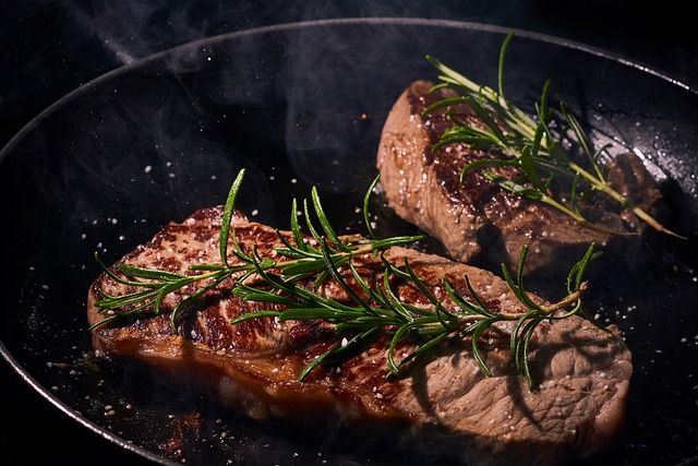 Two steaks sizzling in a pan with rosemary sprigs, steam rising, dark background.