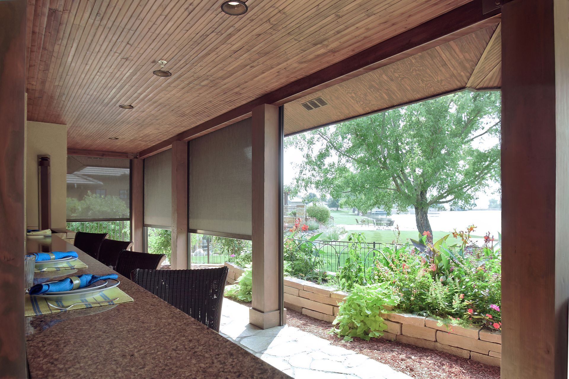 Covered outdoor patio with roll-down shades, looking out to a garden and water.