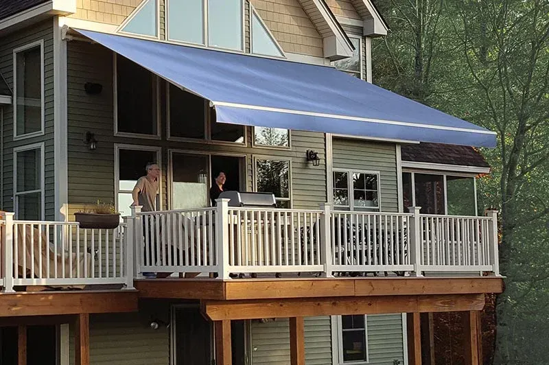 Blue awning shades a wooden deck; a house is in the background, with people visible inside.