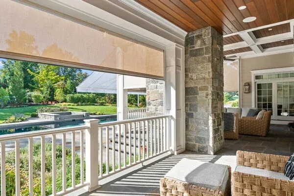 Outdoor patio with pool view; stone pillar and wooden ceiling, retractable shades, wicker furniture.