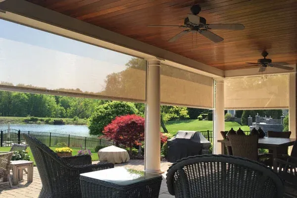 Covered patio with retractable shades, overlooking a lake. Furniture, trees, and two ceiling fans are visible.