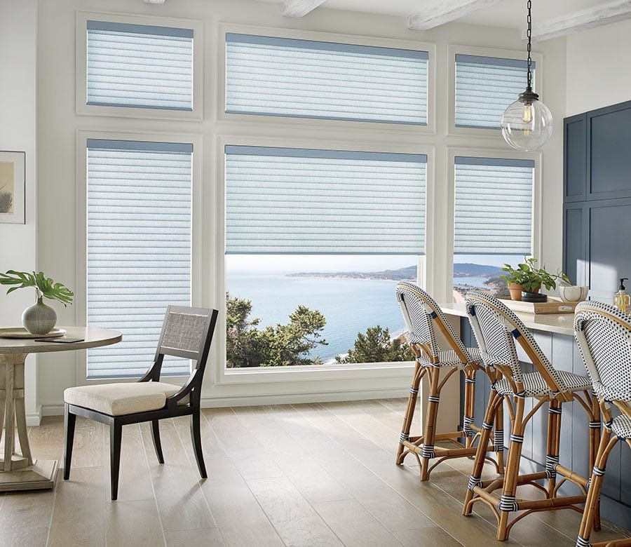 Kitchen with large windows, light blue blinds, and coastal view; rattan bar stools and round table.