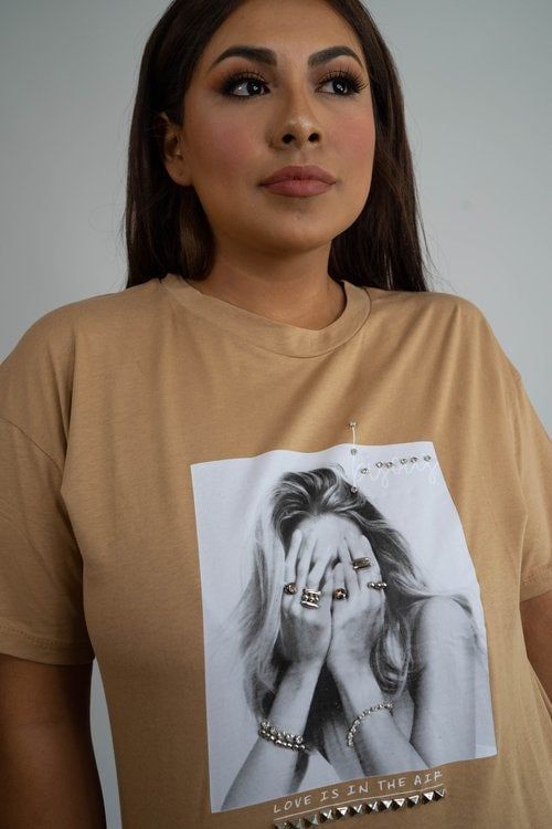 A woman is wearing a tan t-shirt with a picture of a woman covering her face.