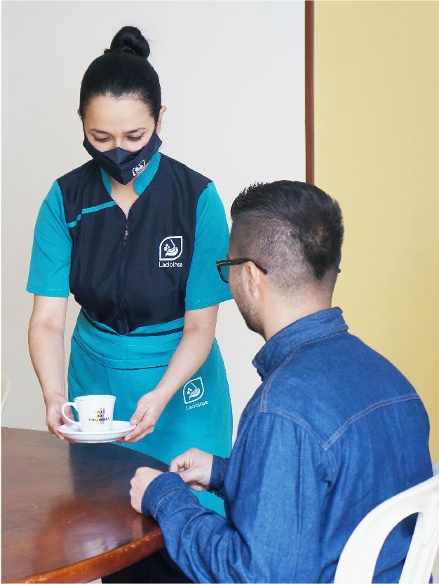 Una mujer con una máscara le sirve una taza de café a un hombre.