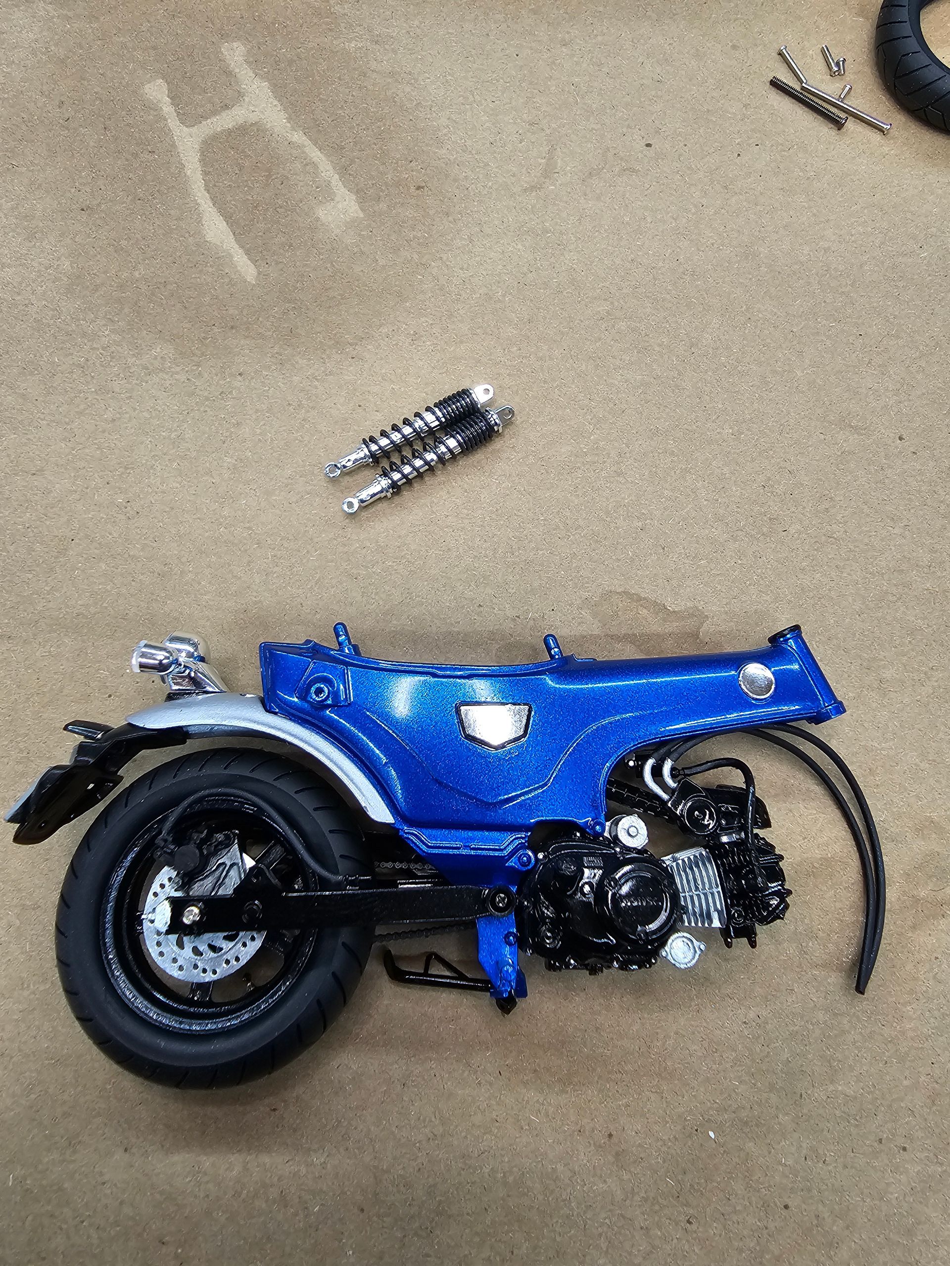 Blue motorcycle model with black engine, rear wheel, and silver exhaust pipes on a textured surface.