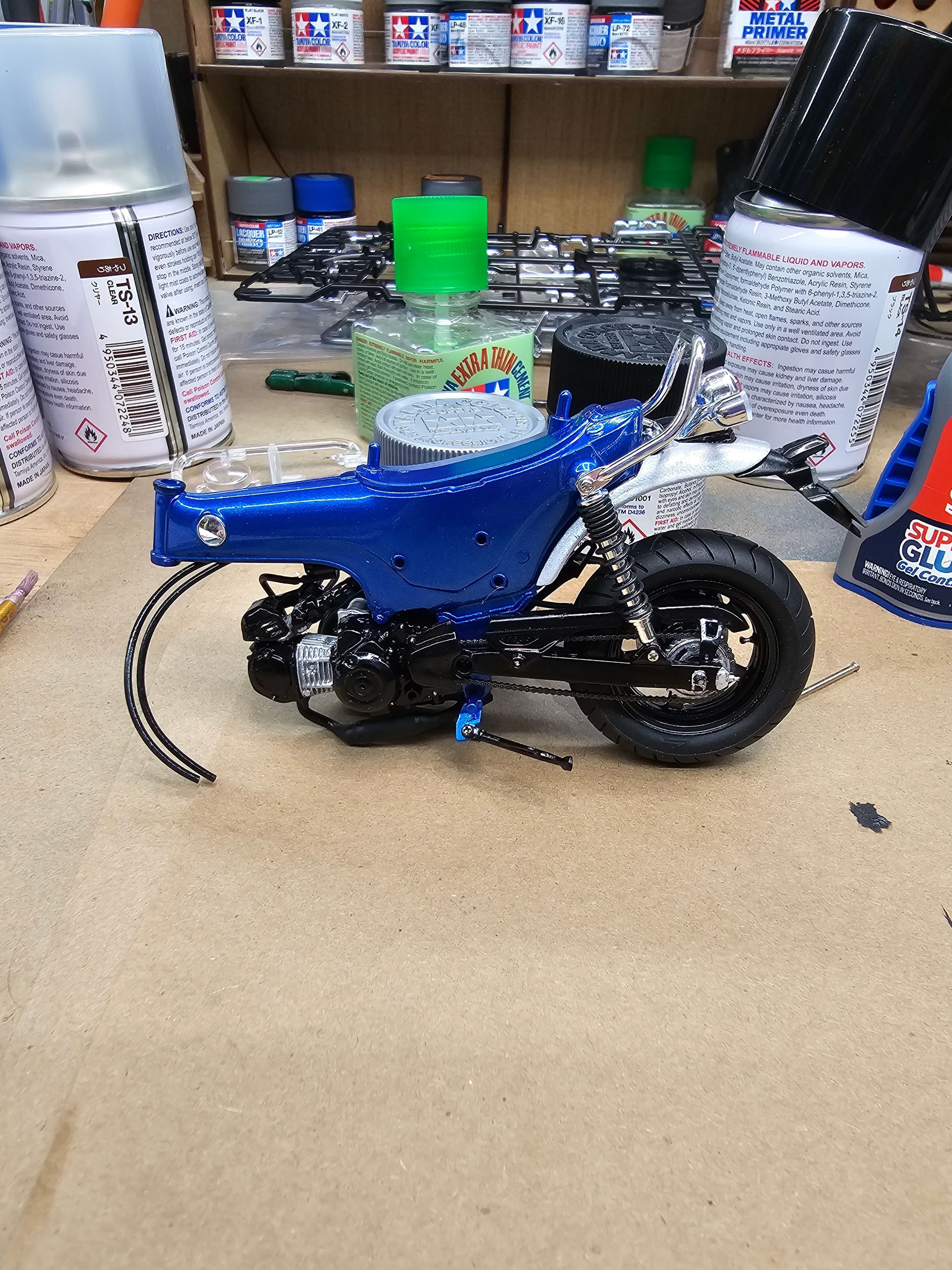 Blue and black model motorcycle on a wooden surface, with paint supplies in background.