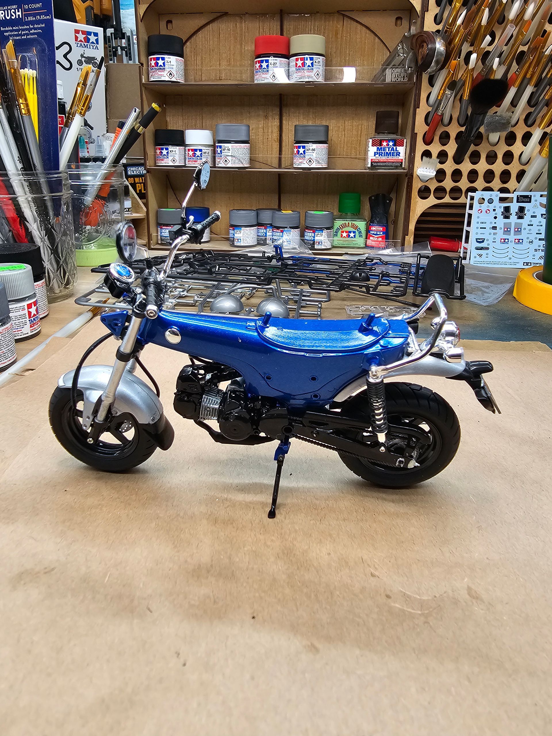 Blue mini-motorcycle in a workshop, parked on the floor, with shelves of paint and tools in the background.