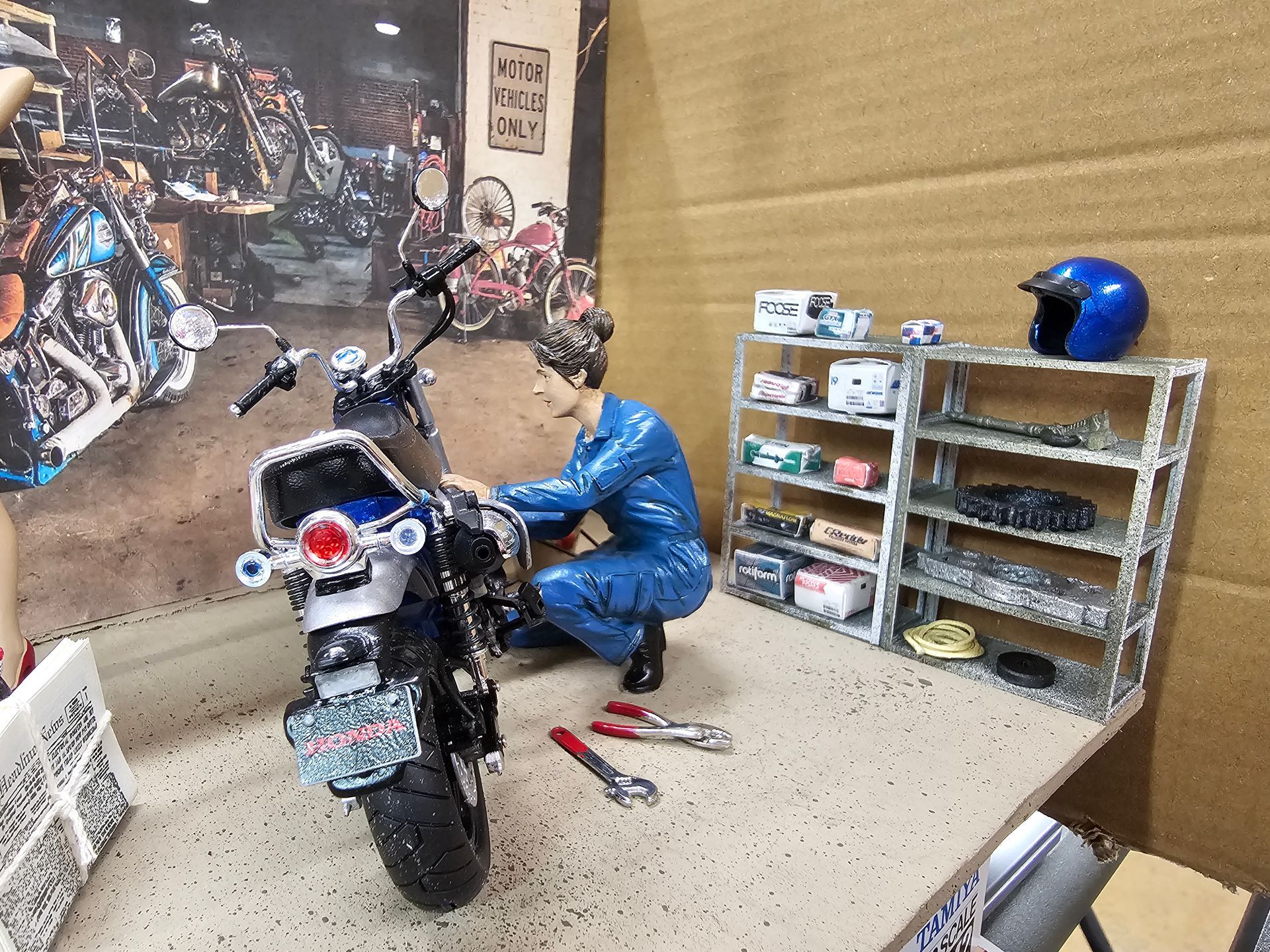 A miniature motorcycle mechanic working on a blue motorcycle in a garage setting with tools and parts shelves.