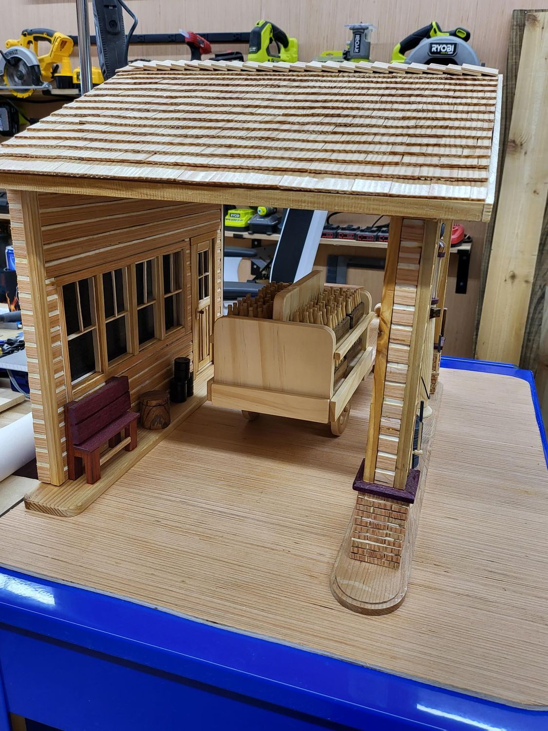 Wooden model of a cabin with a porch swing, shingled roof, and small bench.