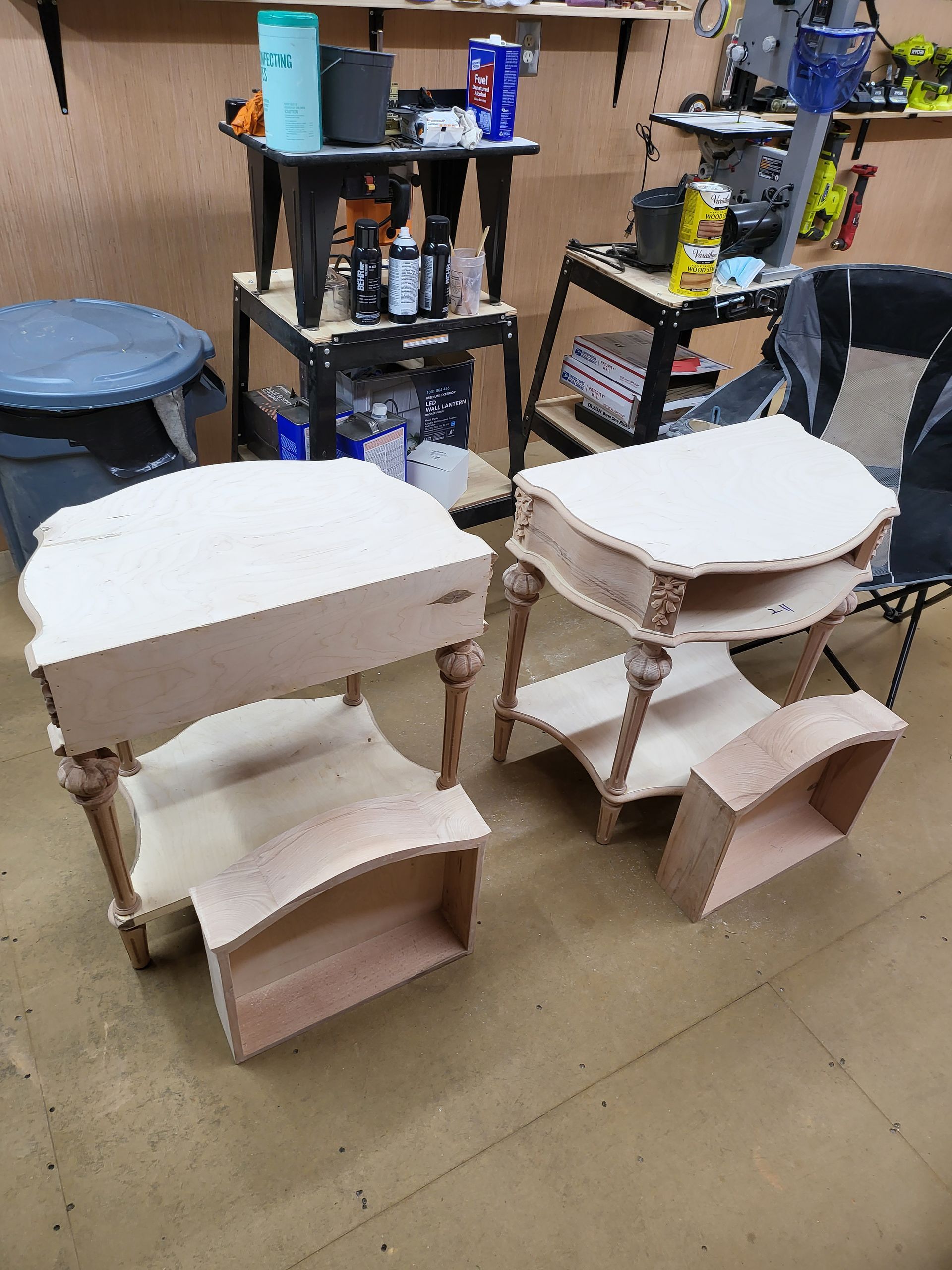 Two unfinished wooden side tables with ornate legs and shelves, in a workshop.