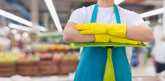 Una donna con un grembiule blu e guanti gialli tiene in mano un vassoio in un supermercato.