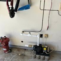 Water pump and plumbing against a white wall in a garage. Tools hang above; wiring and electrical outlets are visible.