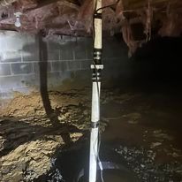 Wooden support post in a crawlspace. Dark, dirt floor. Insulation and cinder block wall visible.