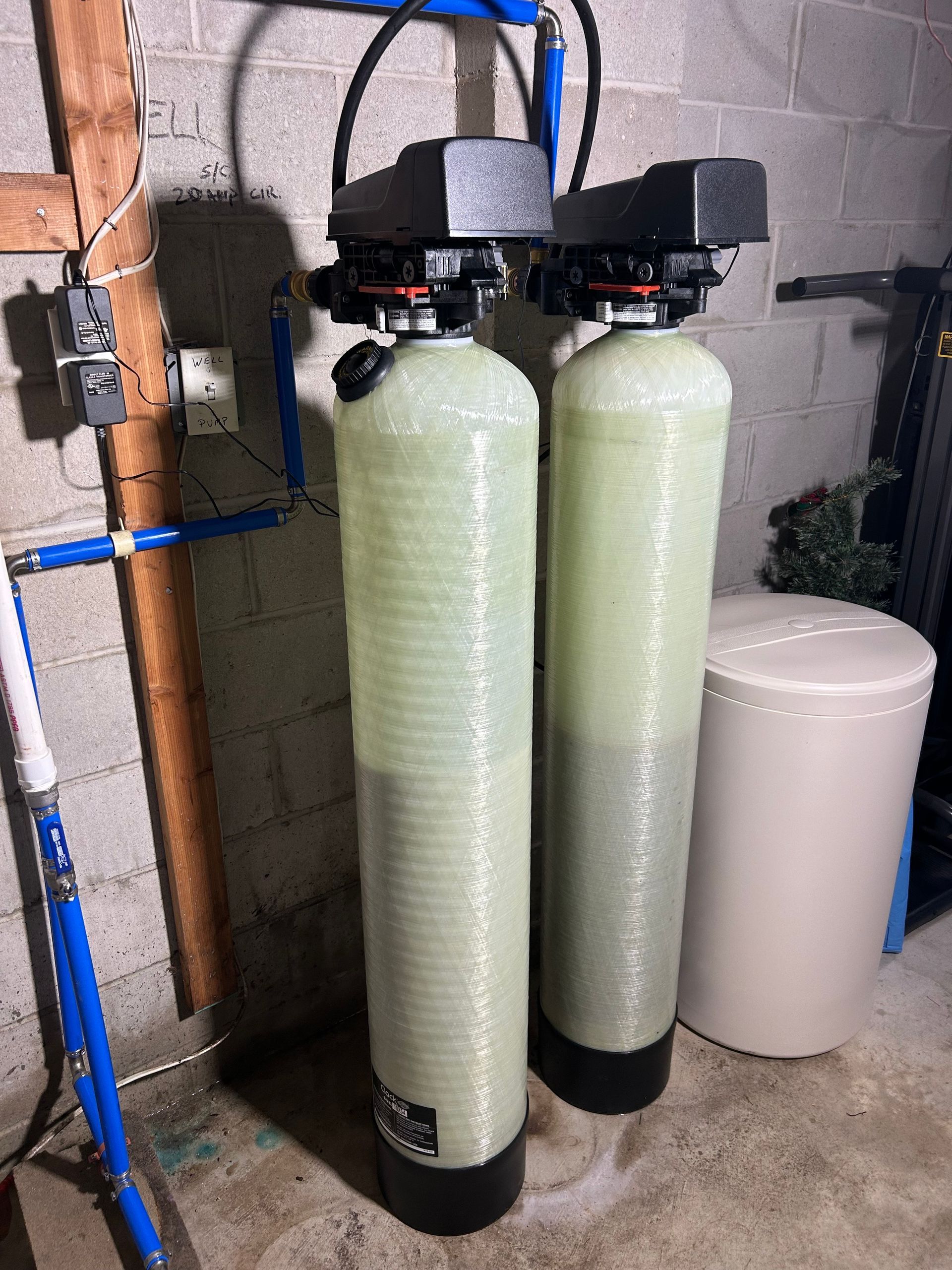 Two tall water filter tanks, blue pipes, and a white tank in a basement.