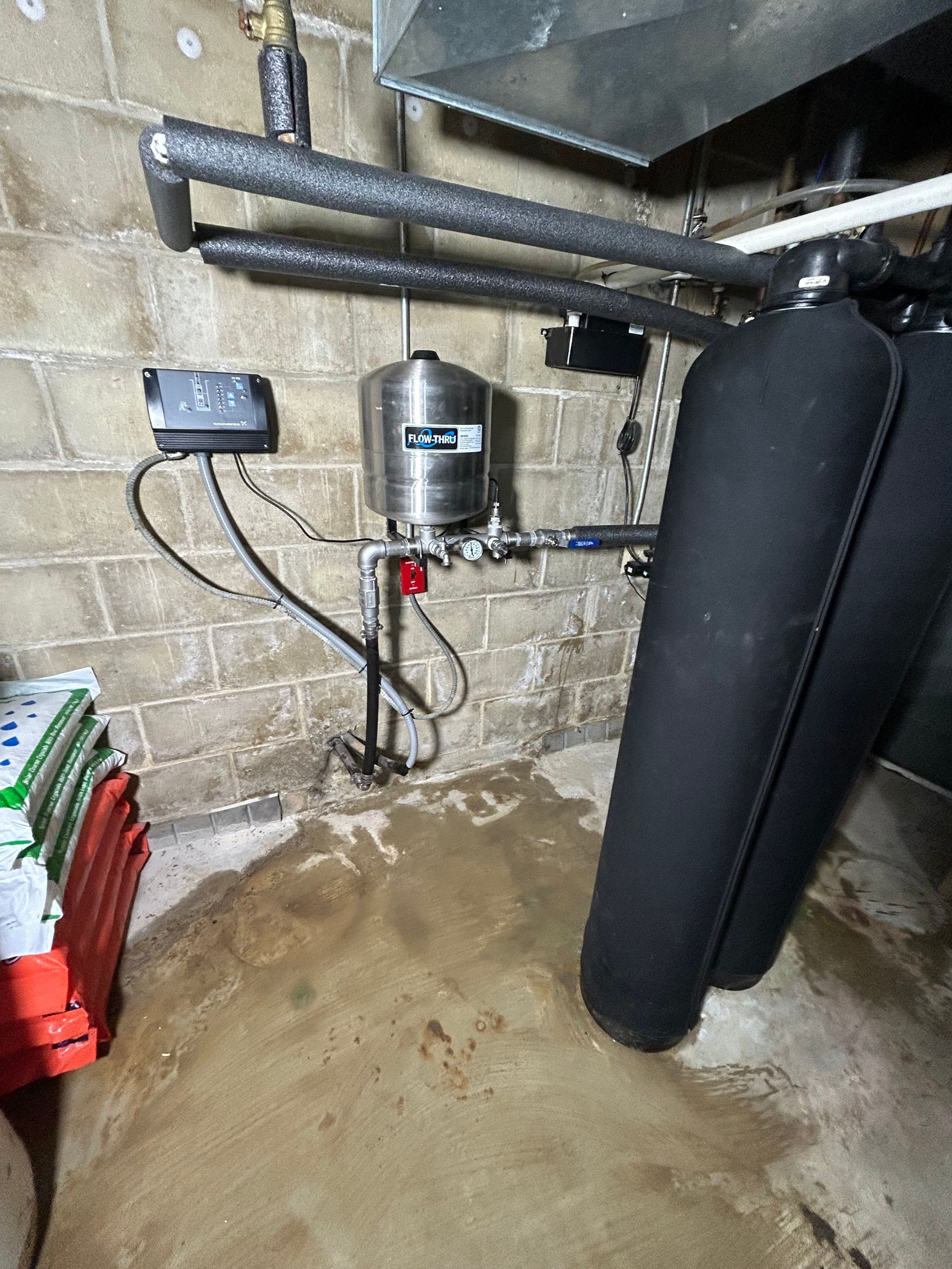 Basement showing a water system with pressure tank, black cylinders, and wet concrete floor.