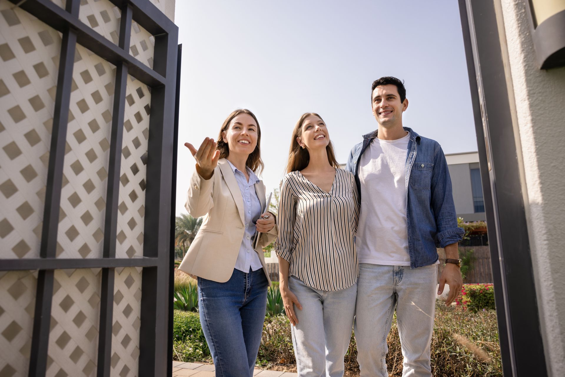 Real estate agent showing house to a couple, all smiling. Open gate, sunny outdoor setting.