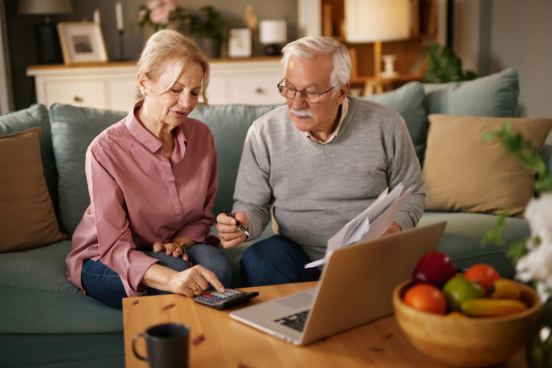 Elderly couple reviews paperwork, using a calculator and laptop, on a living room sofa.