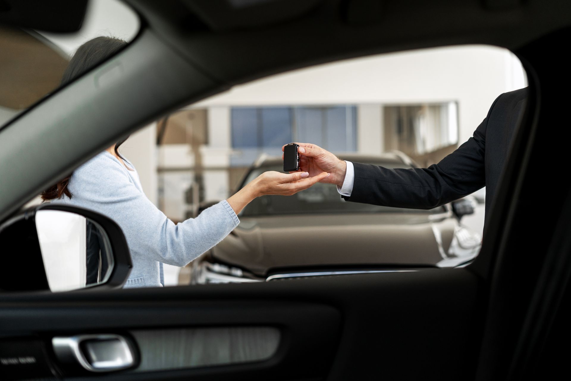 Hand of person in suit gives car keys to person inside the car. Car dealership interior.