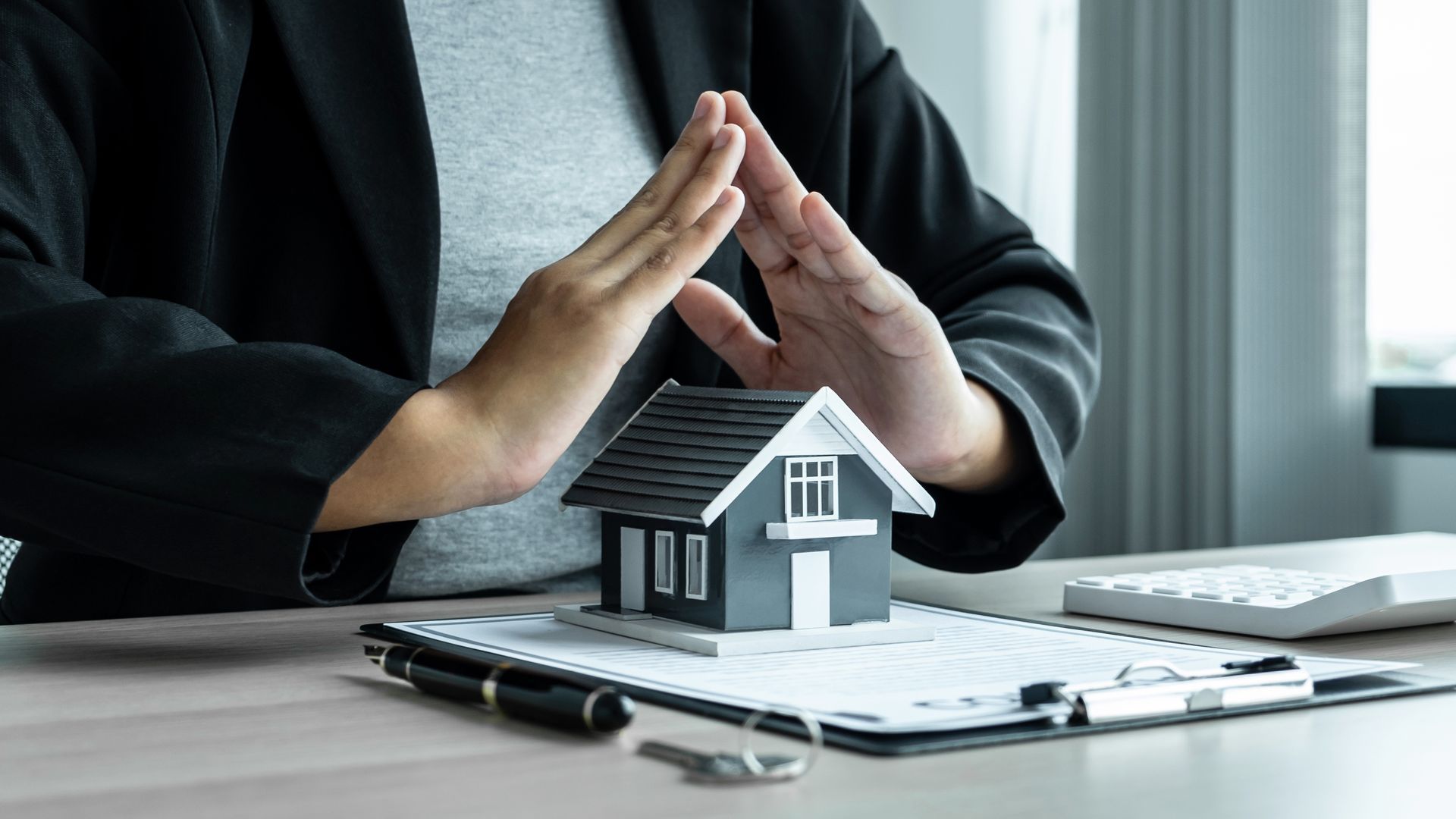 Hands protect a miniature house on a document. Black jacket, pen, and keyboard on a desk.