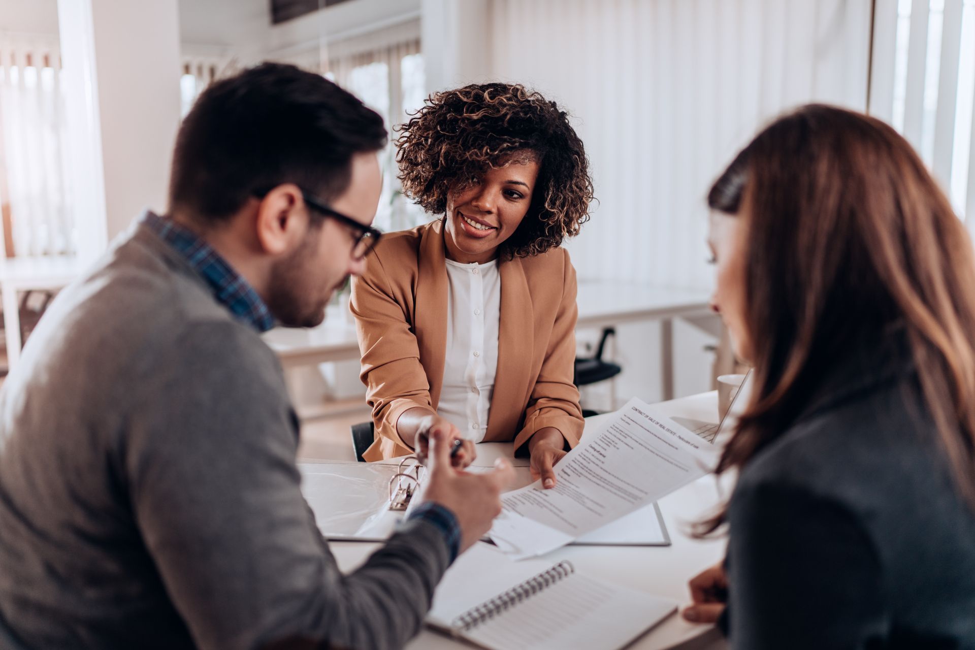 Woman in blazer hands documents to a couple at a desk. Bright office setting.