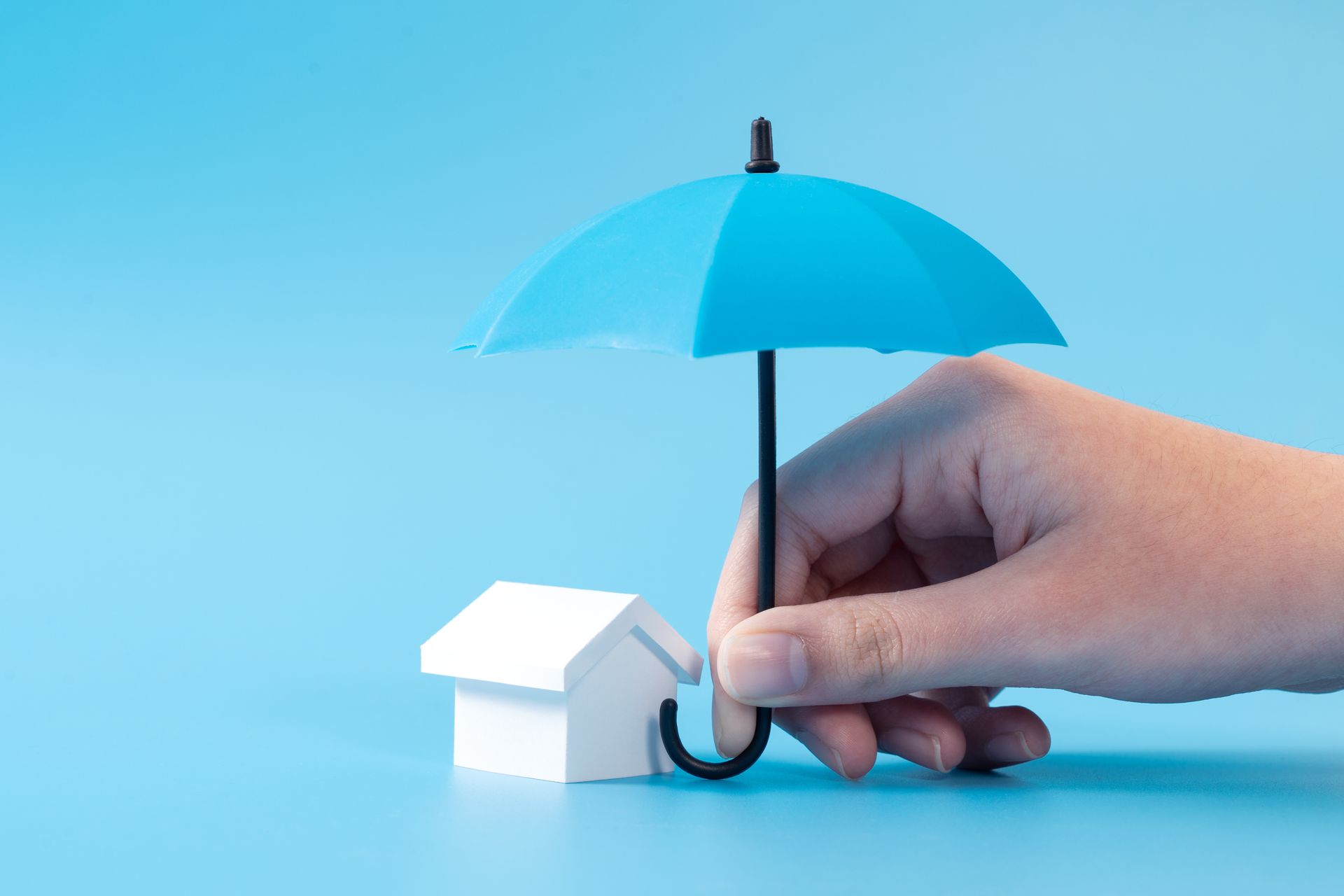 Hand holding a blue umbrella over a miniature white house on a blue background.