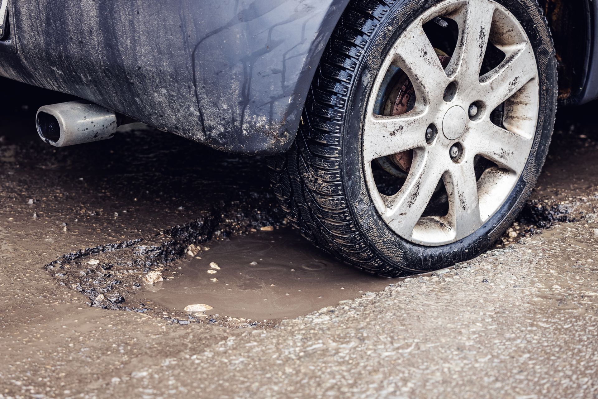 A car tire stuck in a deep pothole filled with water on a damaged and muddy road.