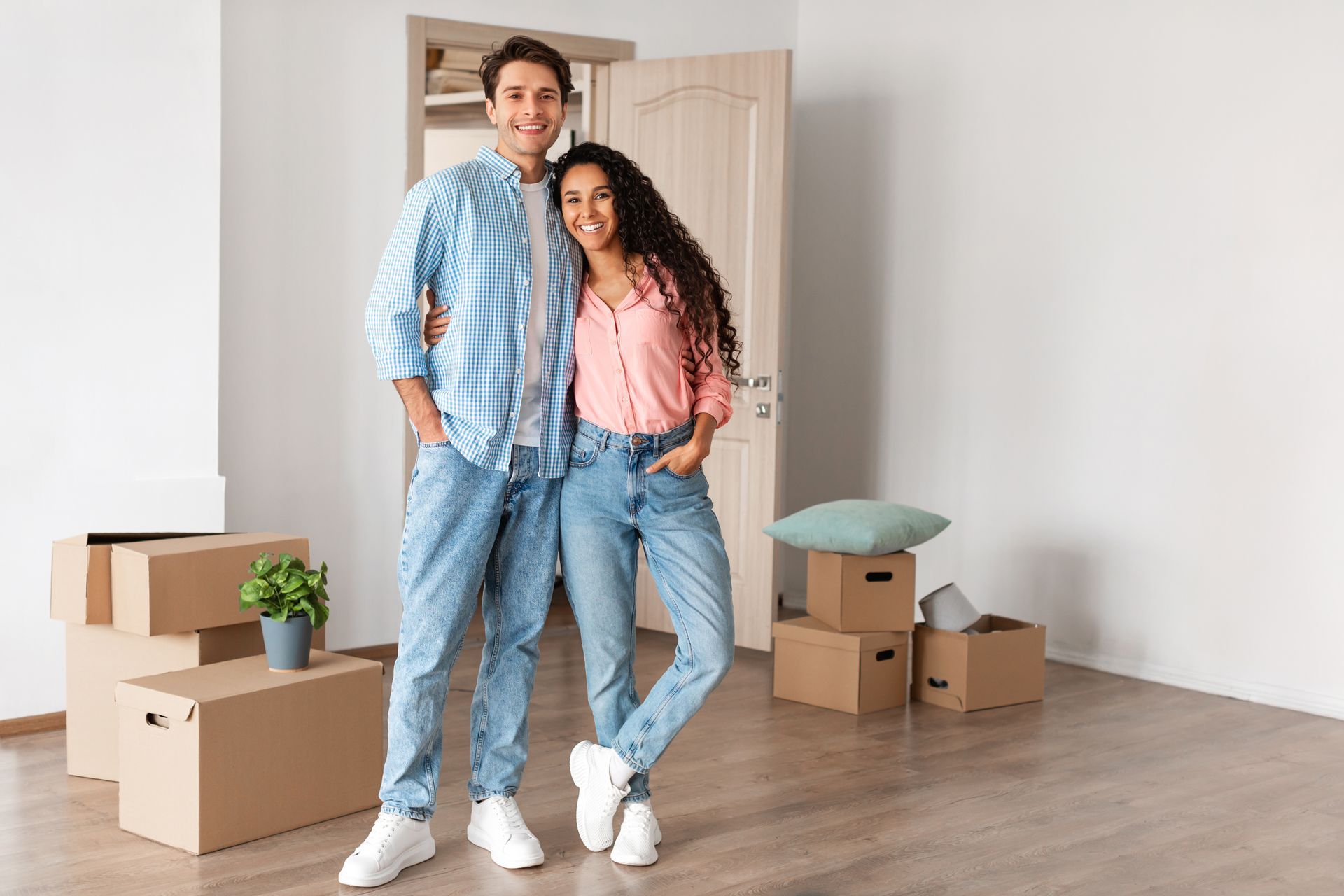 Couple in casual clothing smiles, arms around each other, standing in a new home with moving boxes.