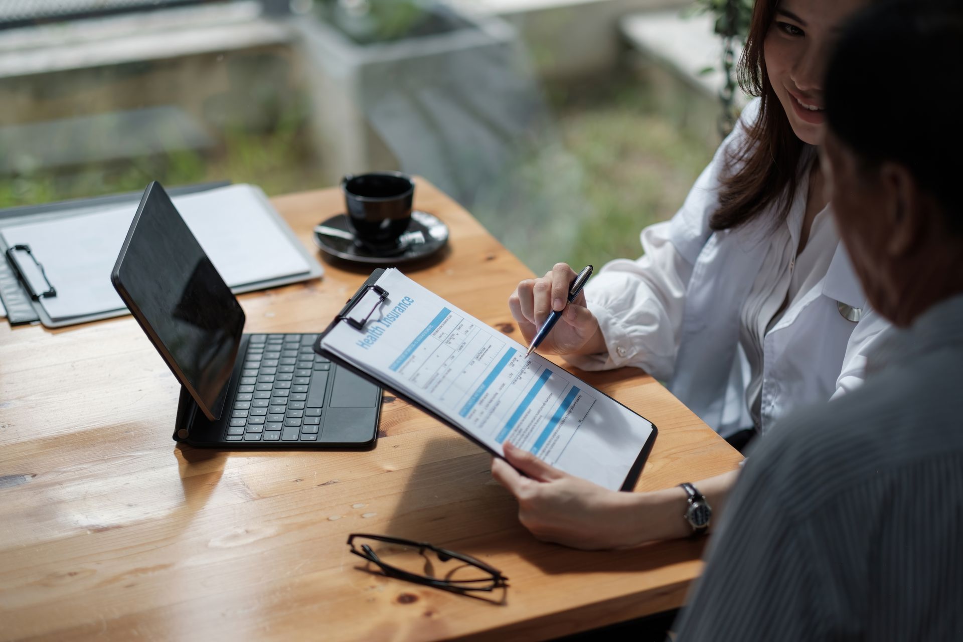 Woman points to document on clipboard, discussing with a person at a table, laptop open, coffee cup, glasses.