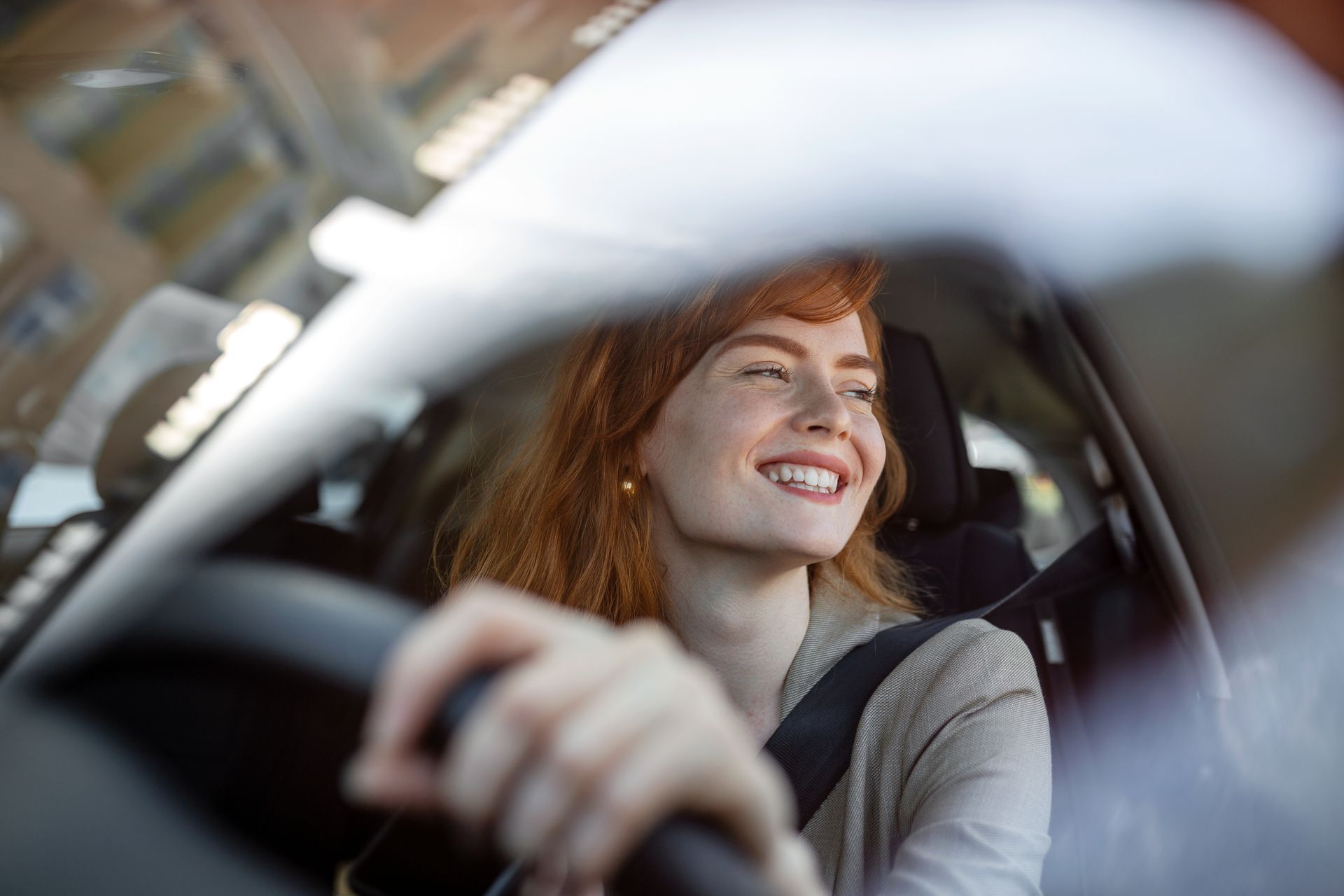 Woman with red hair smiling while driving a car; seatbelt fastened.