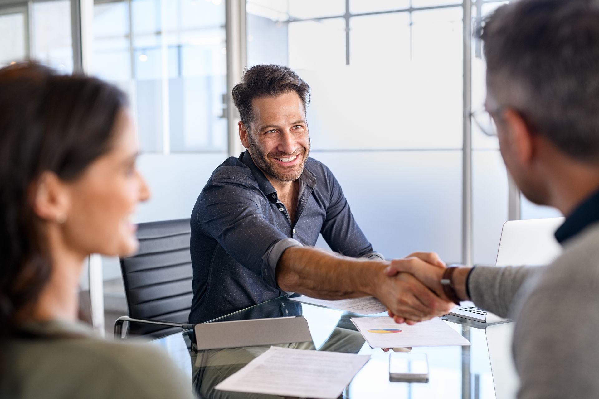 Man shaking hands with another man at a desk; smiling, woman beside them.
