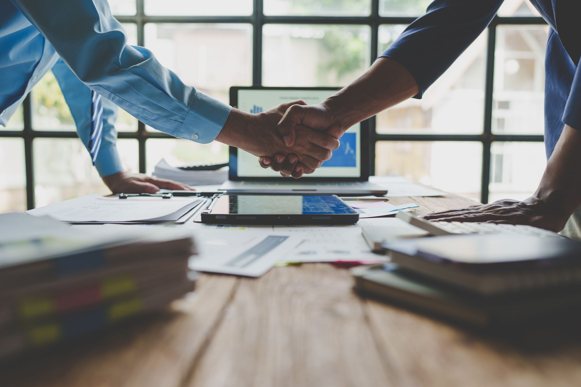 Two people shaking hands over a cluttered desk with a laptop and documents.