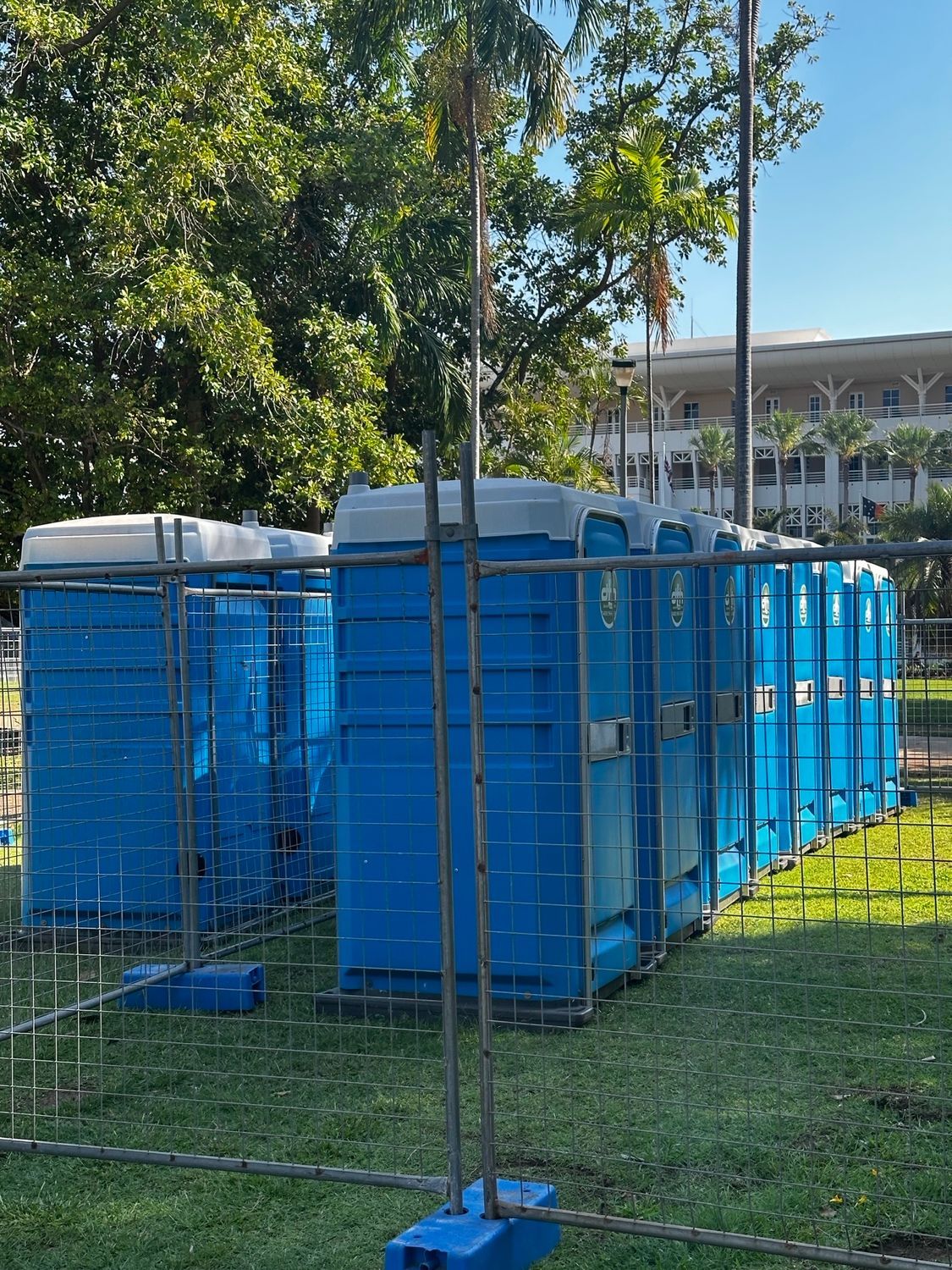 Blue portable toilets behind a chain-link fence on grass with a building and trees in the background - Portable toilet hire for hire in Roseberry, NT
