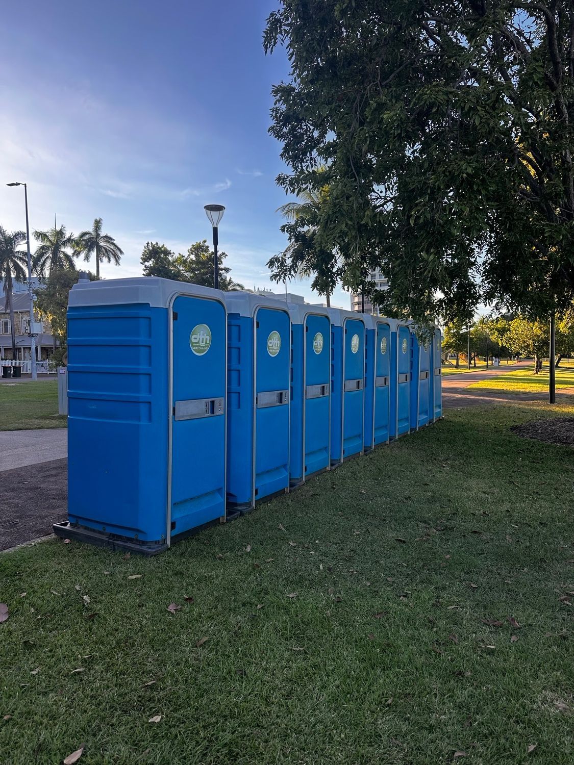 Blue portable toilets lined up on grass in a park, under a blue sky - Portable toilet hire for hire in Roseberry, NT