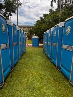 Row of blue portable toilets on green grass; trees and building in the background- Portable toilet hire for hire in Roseberry, NT