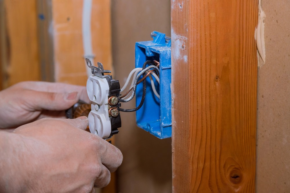 Person installing an electrical outlet with exposed wires and a blue electrical box in a wooden wall.