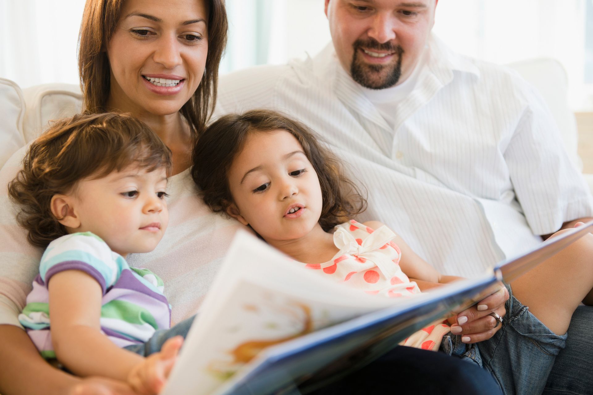 Family reading a book together on a couch; mother and father with two young daughters.