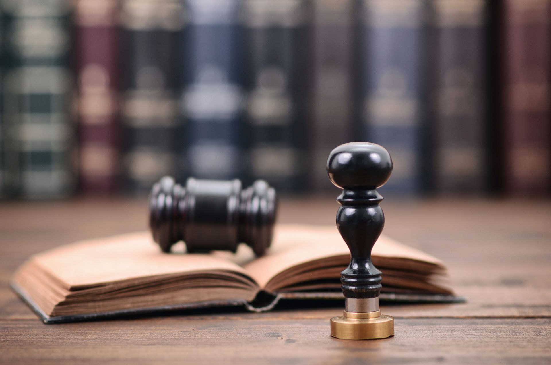 Gavel and seal resting on an open book, against a blurred background of bookshelves.
