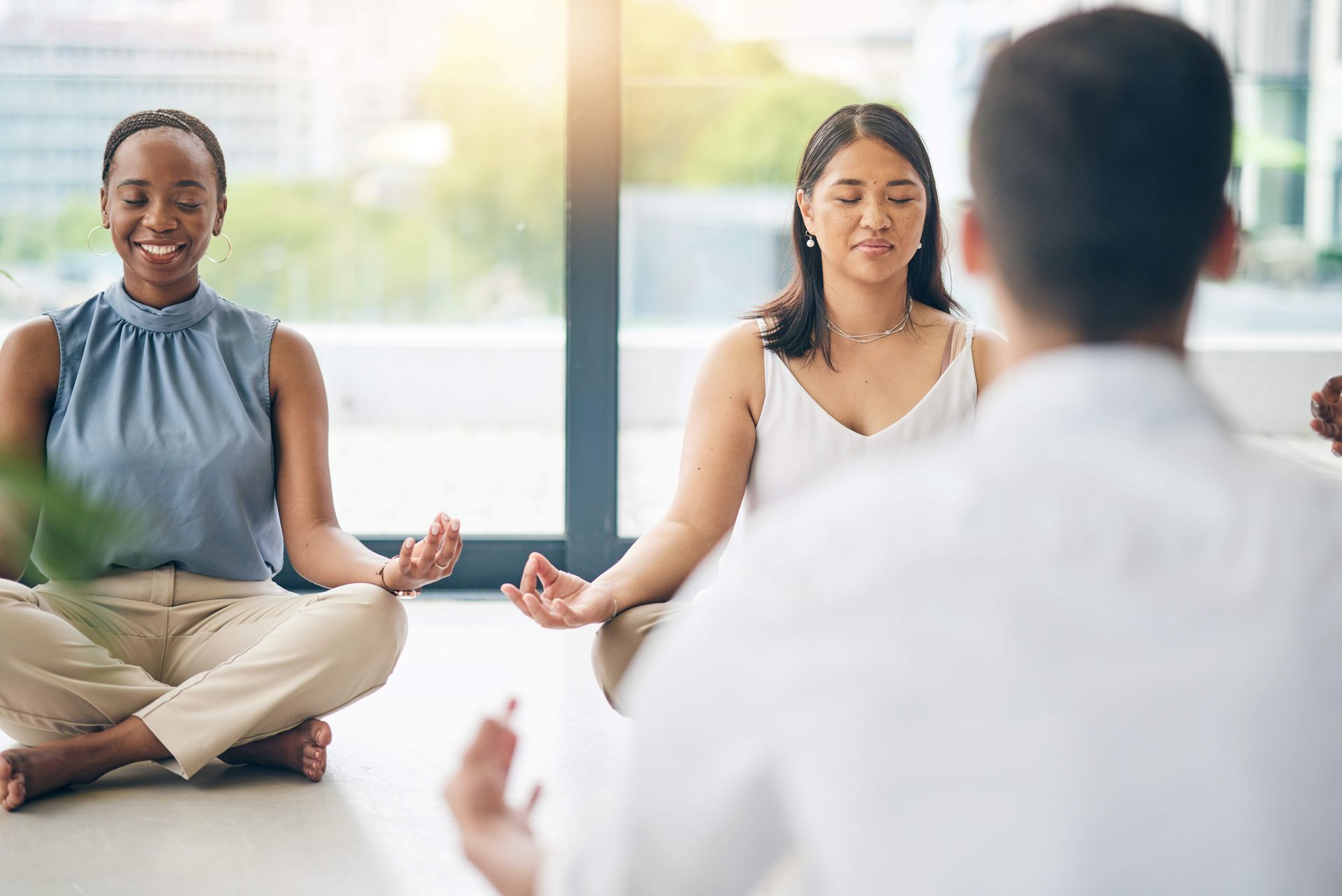 Three people sit cross-legged in a bright room, meditating during a group session.