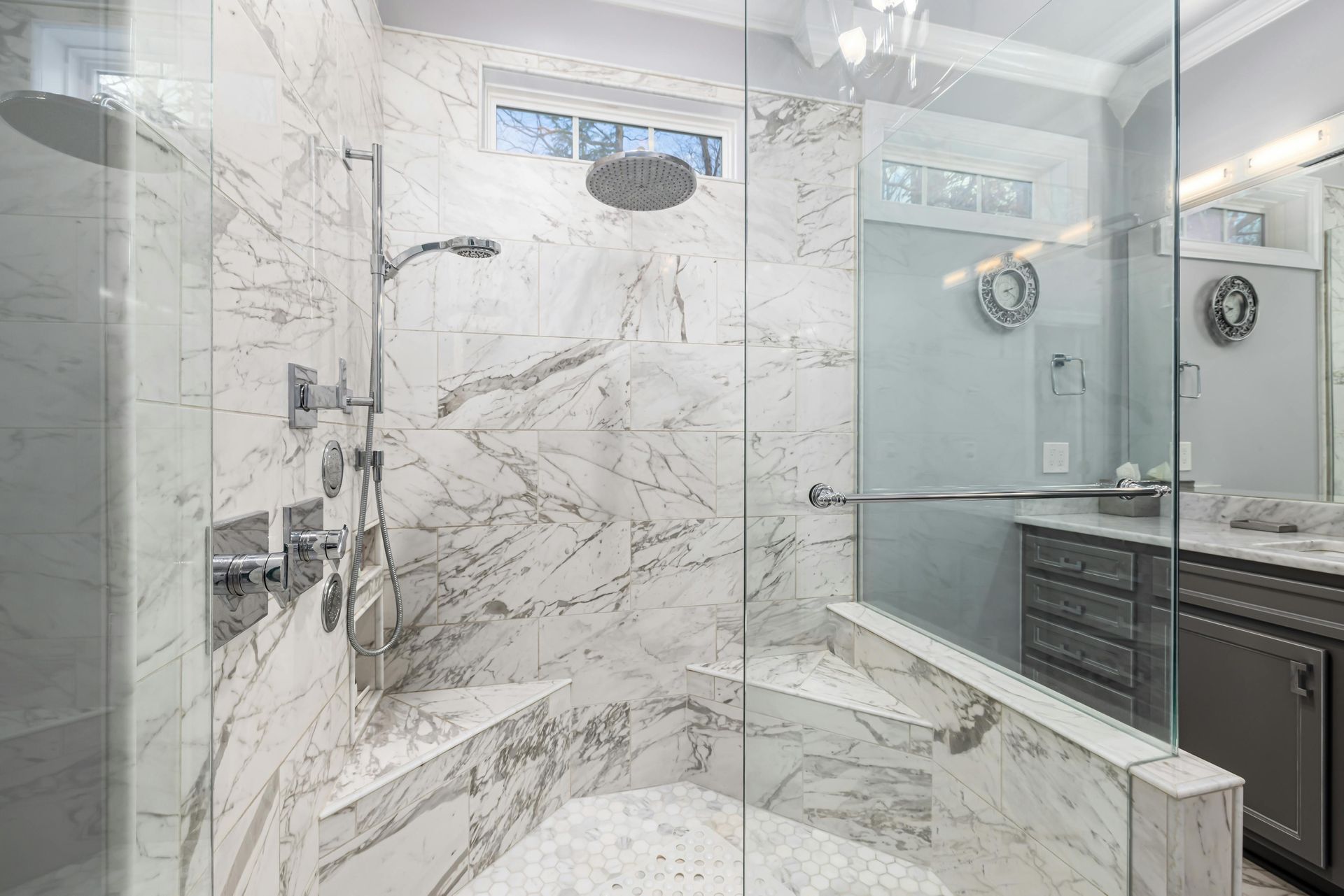 A luxury walk-in shower with white marble walls, two corner benches, a rainfall showerhead, and glass enclosure panels.