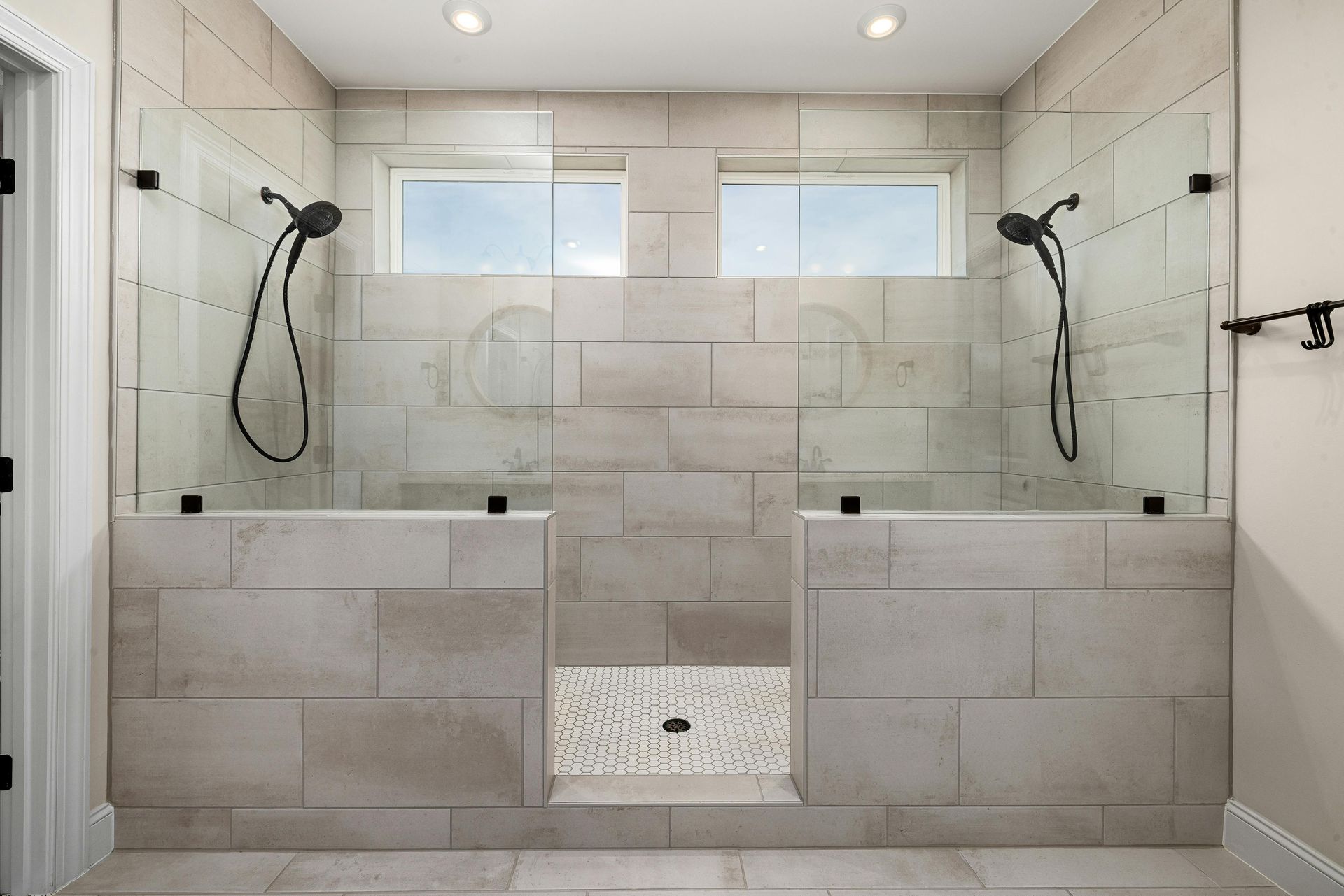 A modern walk-in shower with light stone tile walls, two glass panels, dual shower heads, and a pebbled floor.