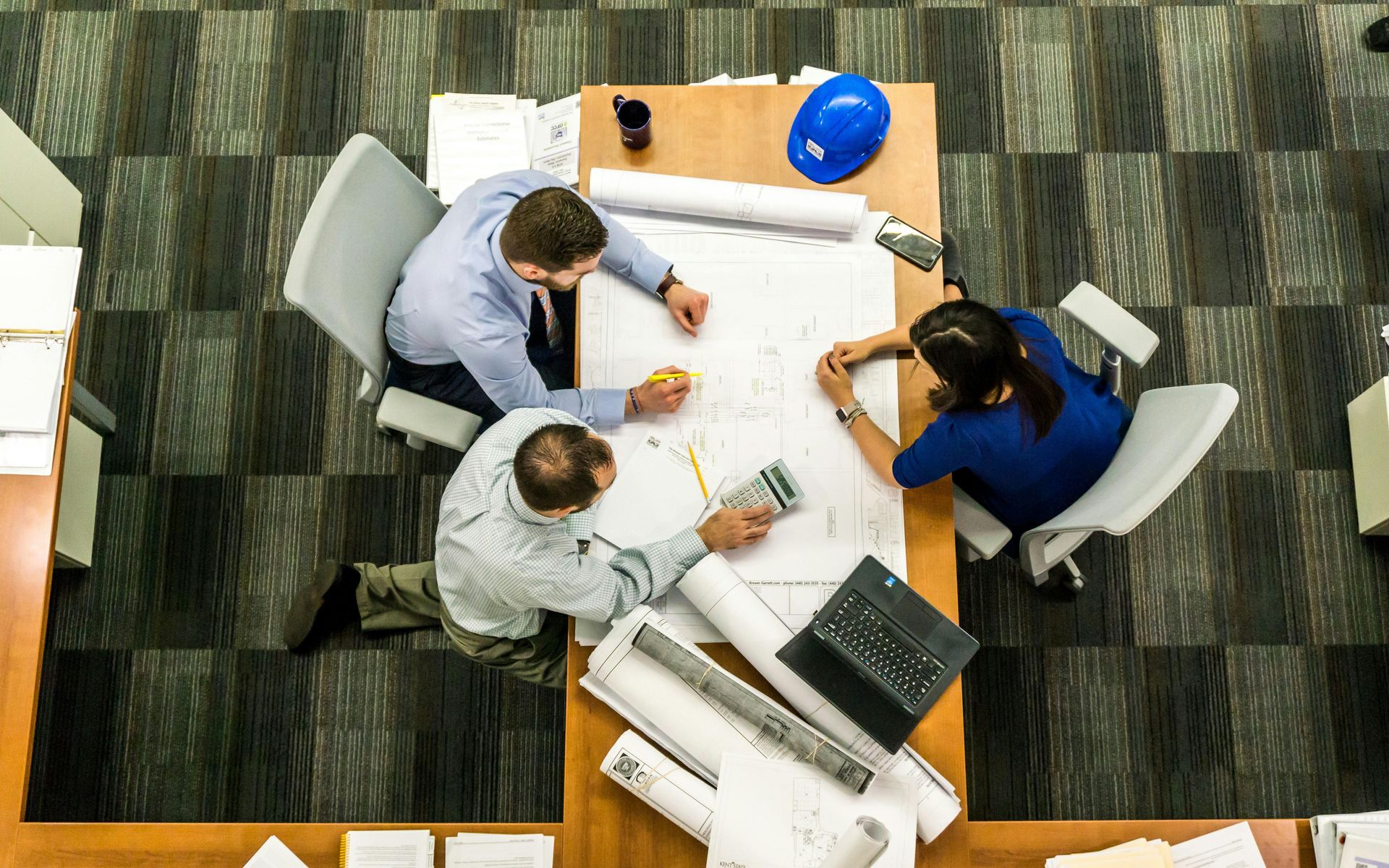 Three people at a table reviewing blueprints, one has a hard hat.