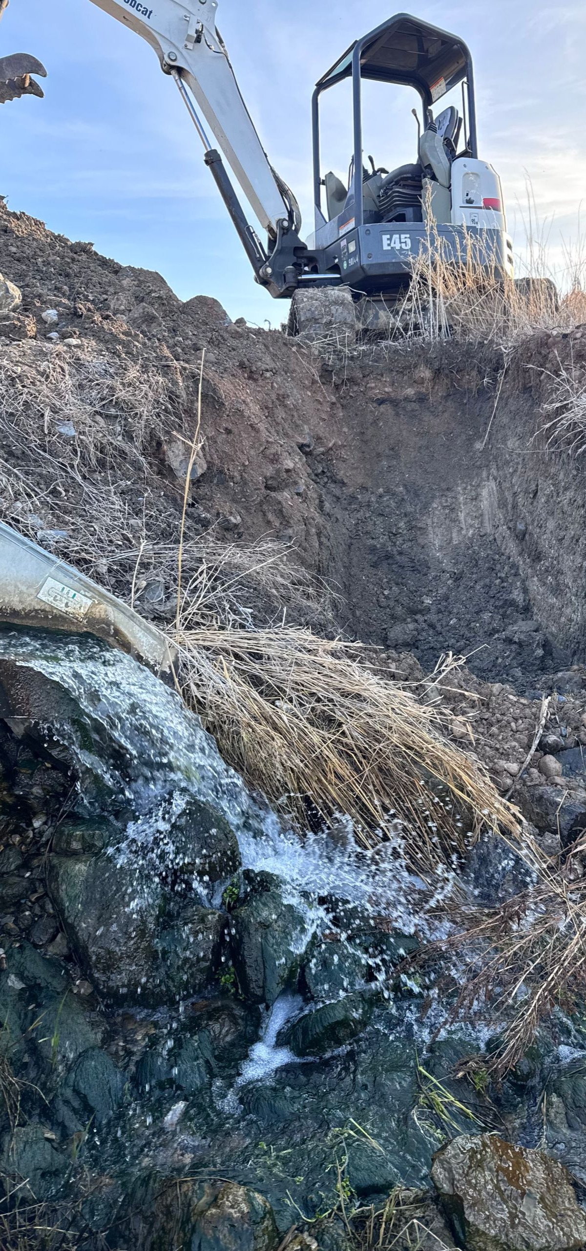 An excavator digs into a muddy hillside. White substances are visible on the soil. Blue sky in the background.