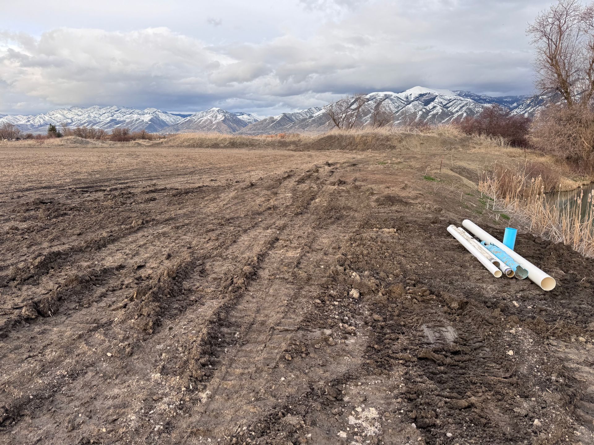 Muddy dirt field with white pipes, snow-capped mountains in the background under cloudy sky.