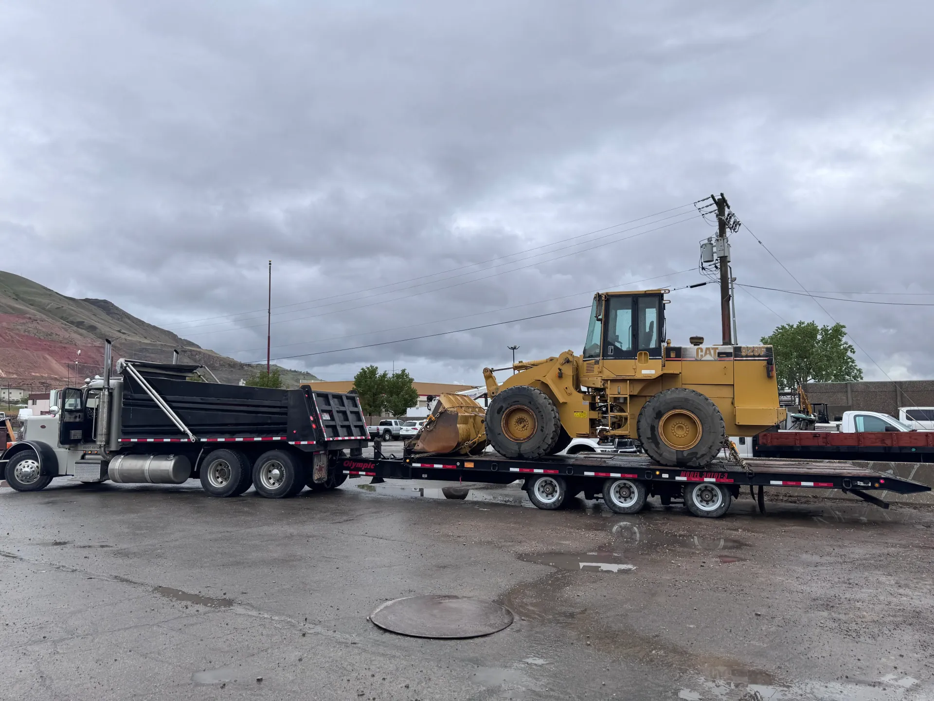 Truck hauling a large yellow construction loader on a trailer; overcast sky.