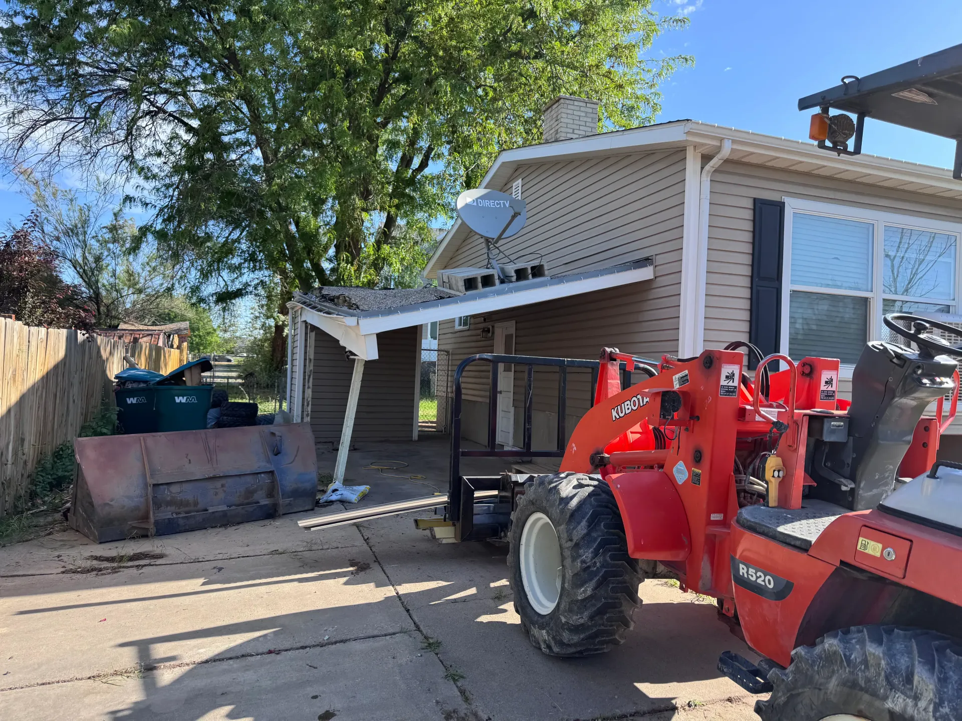A red tractor is parked next to a small beige house with a detached awning and satellite dish.